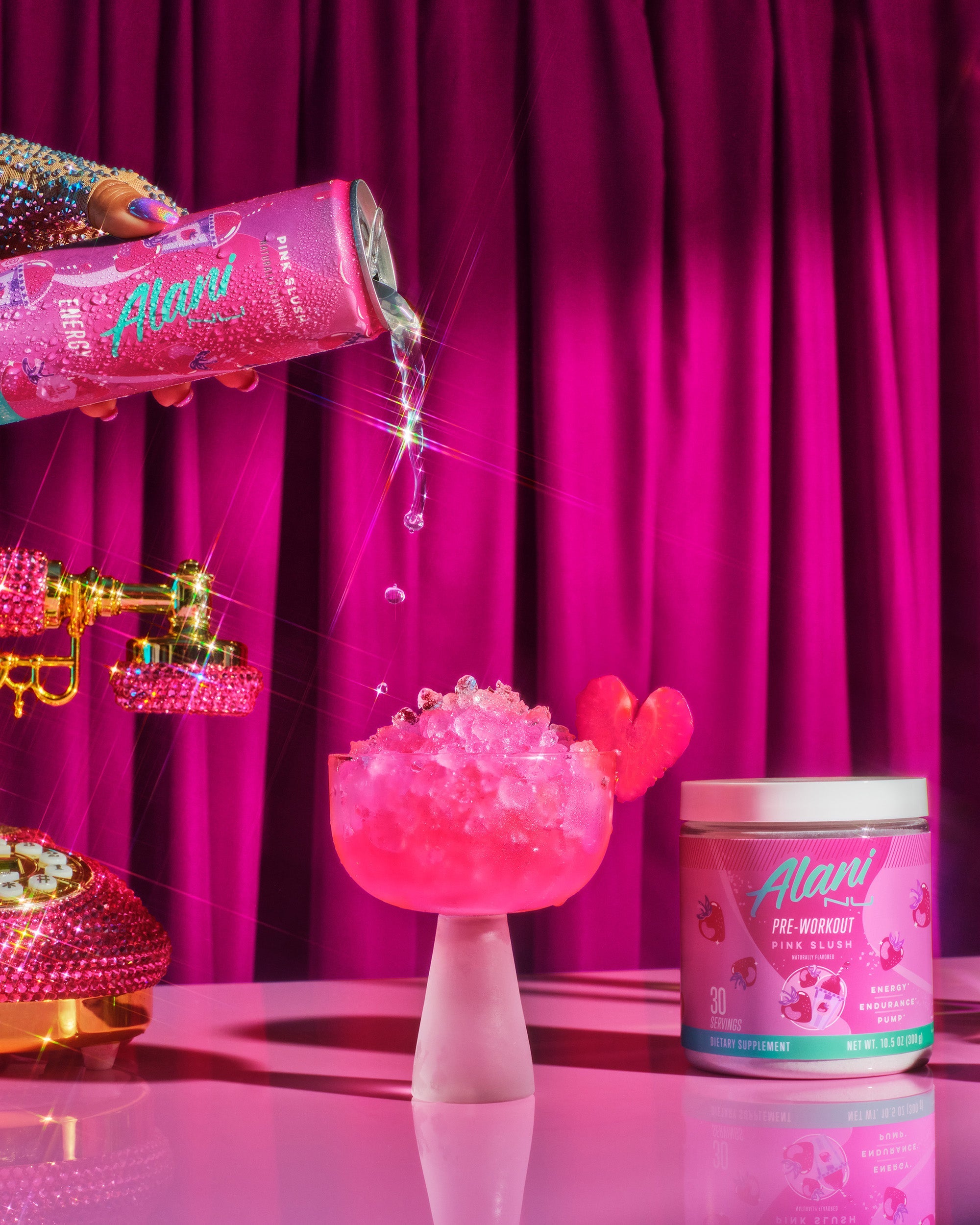 A can of Pink Slush Energy pouring into a frosted coup glass filled with pebble ice and pink liquid. Pink Slush Pre-Workout and a bedazzled pink phone are on either side of the glass.